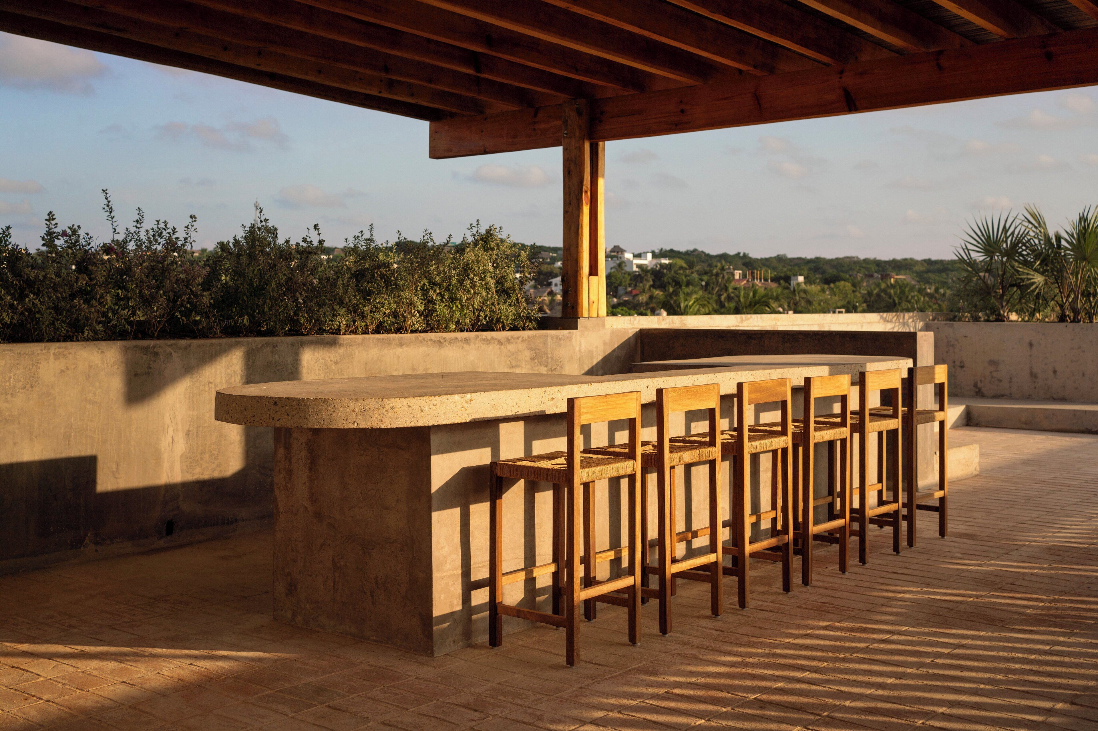 Un área de bar al aire libre con mostrador de hormigón y sillas de madera, con un fondo de follaje y edificios.