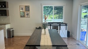 Un espace repas avec une longue table, un piétement blanc et un plateau foncé. Des œuvres d’art encadrées ornent le mur, une fenêtre offre une vue sur la verdure et une poubelle est à disposition.
