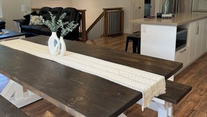 Une cuisine moderne avec un réfrigérateur en acier inoxydable, une table à manger en bois agrémentée d’un chemin de table blanc, et un coin salon avec un canapé en cuir foncé et un vase rempli de verdure.
