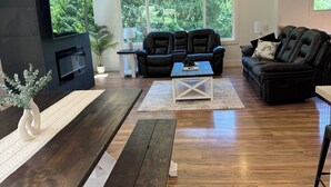 Un salon moderne doté d’une cheminée, d’un parquet en bois foncé, d’un canapé en cuir noir et d’une grande fenêtre donnant sur un paysage verdoyant.