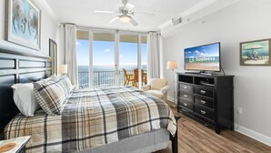 Habitación de hotel con una cama grande, un televisor, un balcón con vistas al mar y un ventilador de techo.