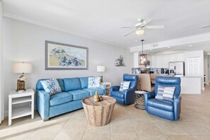 A spacious living room with a blue sofa, a leather armchair, and a round coffee table. A ceiling fan and a kitchen area are visible in the background.