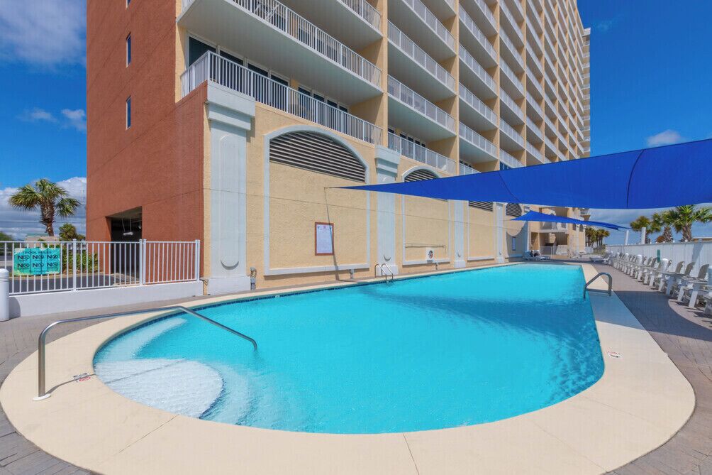 A swimming pool with a curved edge, a building with multiple balconies, and a clear blue sky.