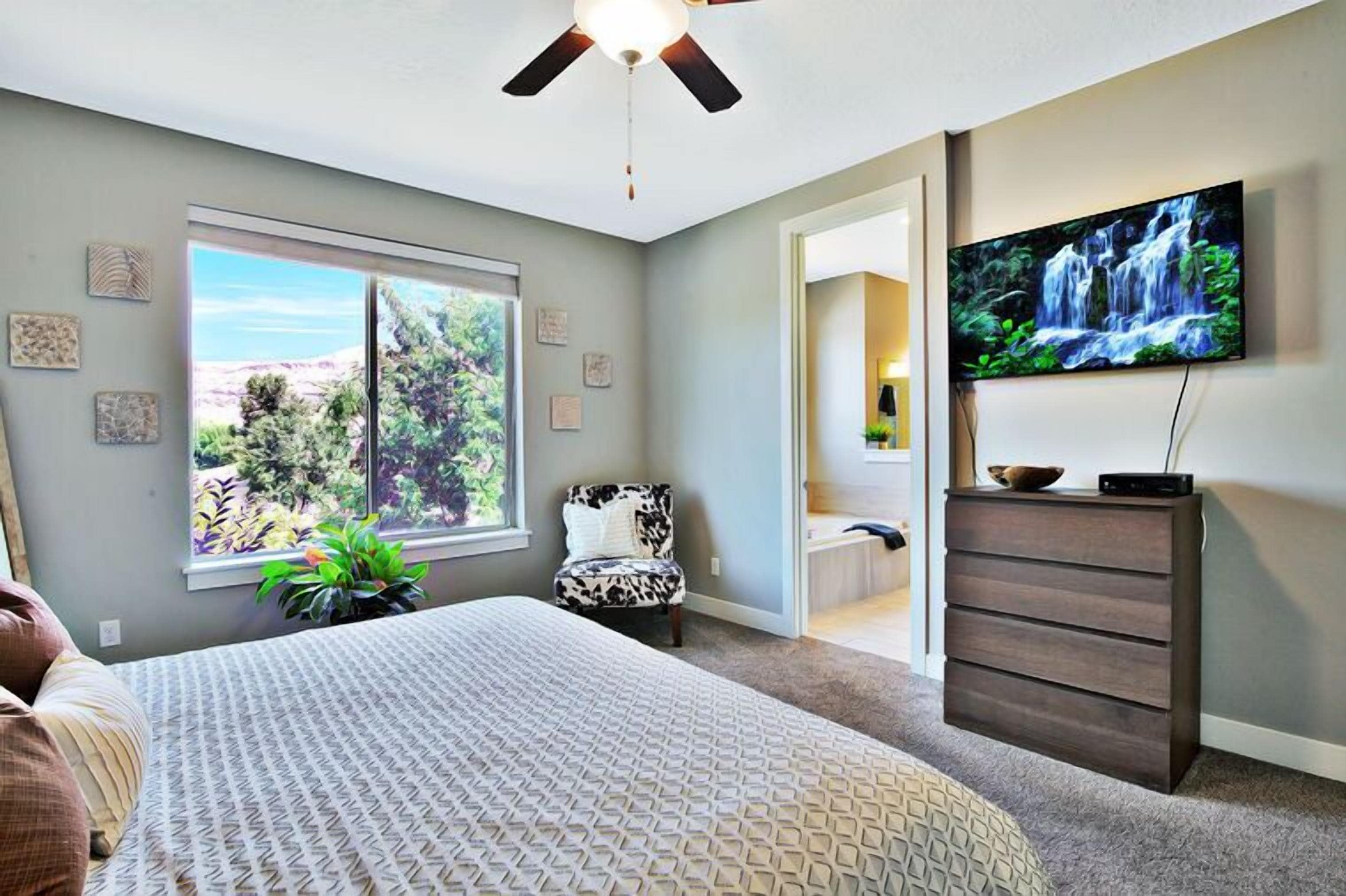 A bedroom with a bed, a ceiling fan, a dresser, a TV, and a window with a view of trees and a mountain.