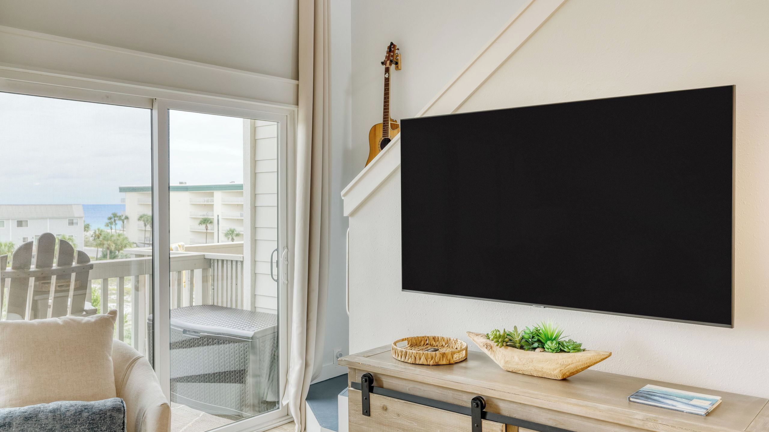 A balcony with a view of buildings and the ocean, a guitar, a blackboard, and a wooden table with a plant and a basket.