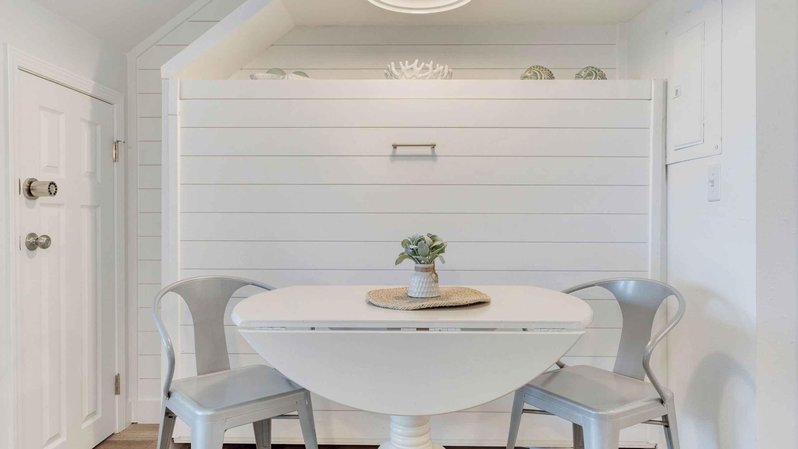 Een eethoek met een ronde tafel, twee stoelen, een kleine plant en een wandplank met decoratieve objecten.
