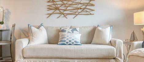 A living room with a beige sofa, decorative pillows, a wooden coffee table with seashells, and a wall art piece.