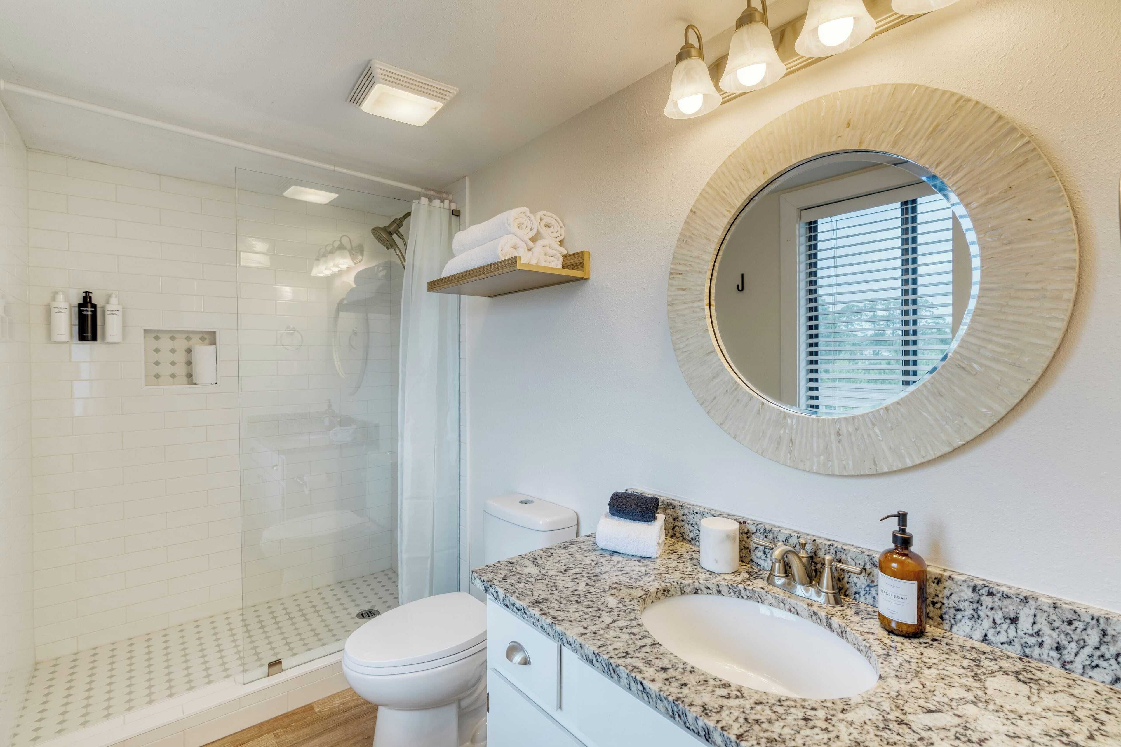 A modern bathroom with a glass-enclosed shower, a white toilet, a granite countertop, a round mirror, and a shelf with toiletries.