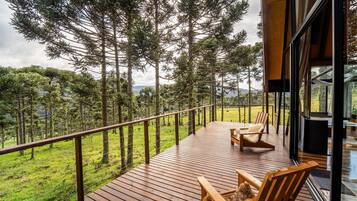 A wooden deck with two wooden chairs, overlooking a forested area with tall trees and a grassy field.