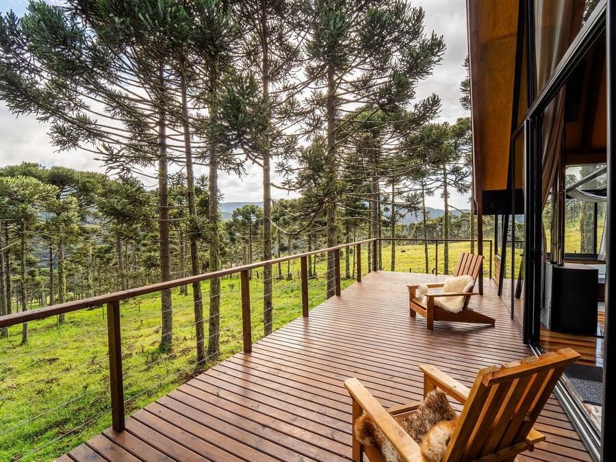 A wooden deck with two wooden chairs, overlooking a forested area with tall trees and a grassy field.