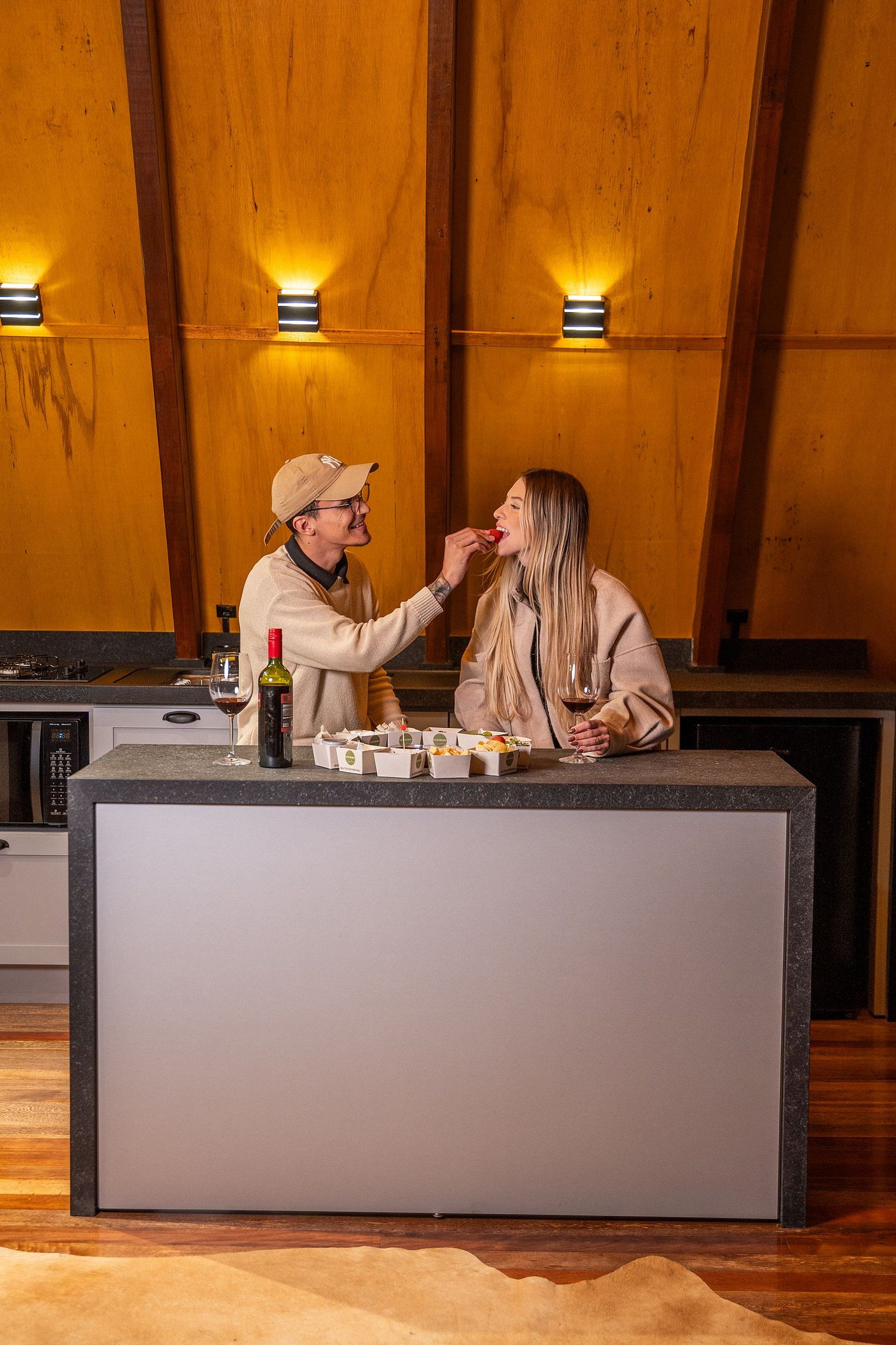 Una cocina moderna con paneles de madera, una isla central con una botella de vino y copas, y una persona sirviendo comida.