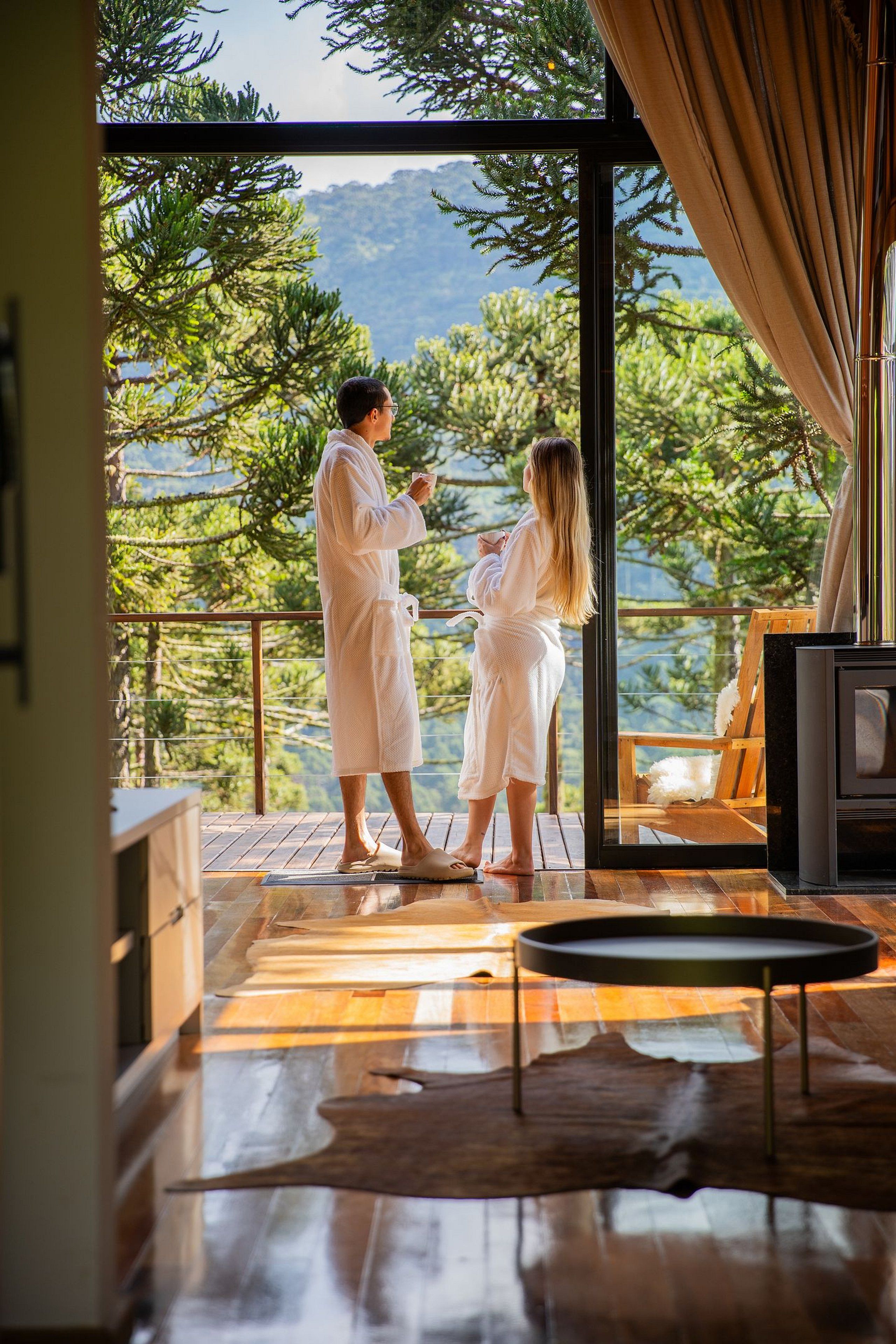 A couple in white bathrobes standing by a glass door, overlooking a mountainous landscape.