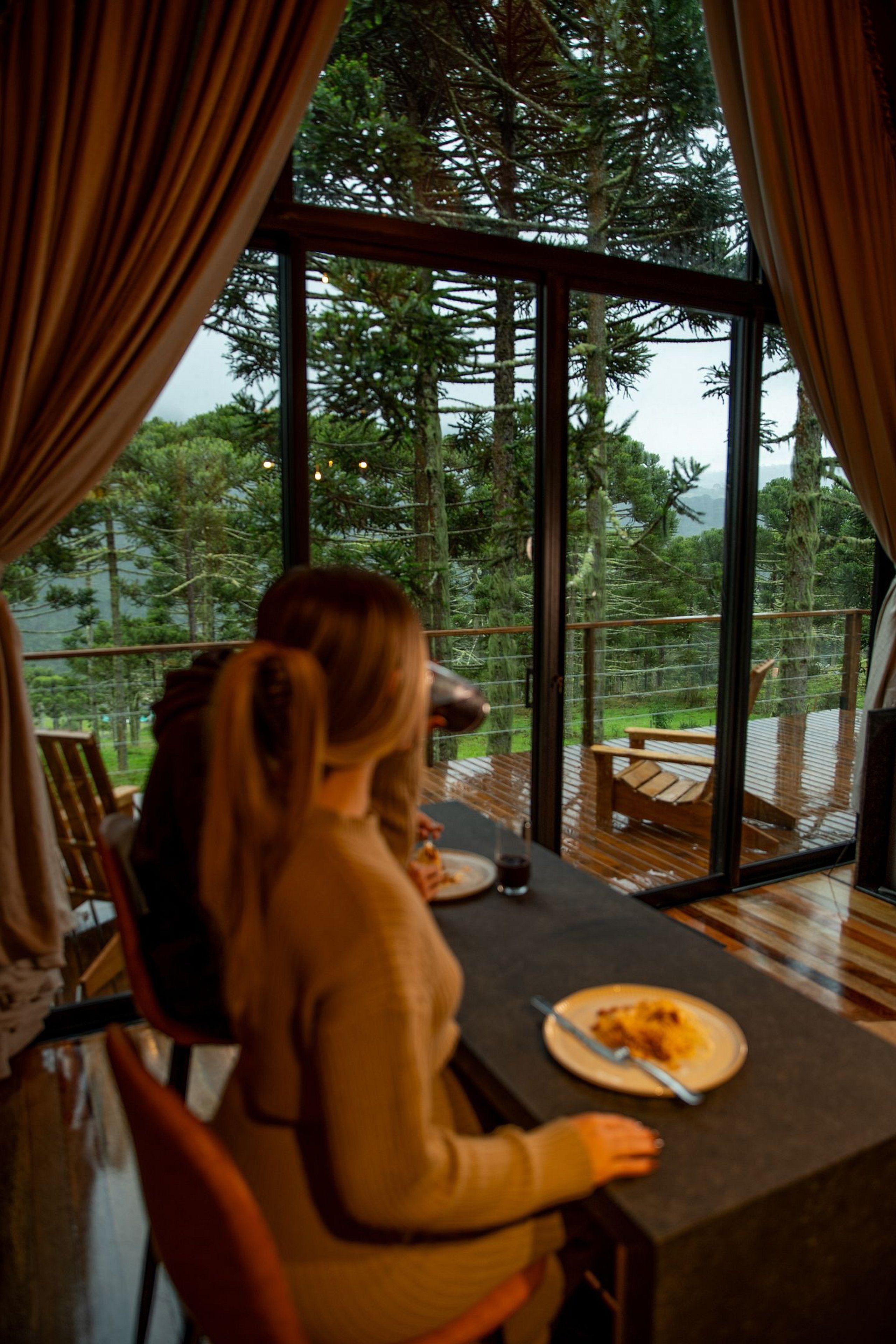 Uma área de refeições com mesa e cadeiras, um prato de comida e vista para um deck com assentos externos e árvores.