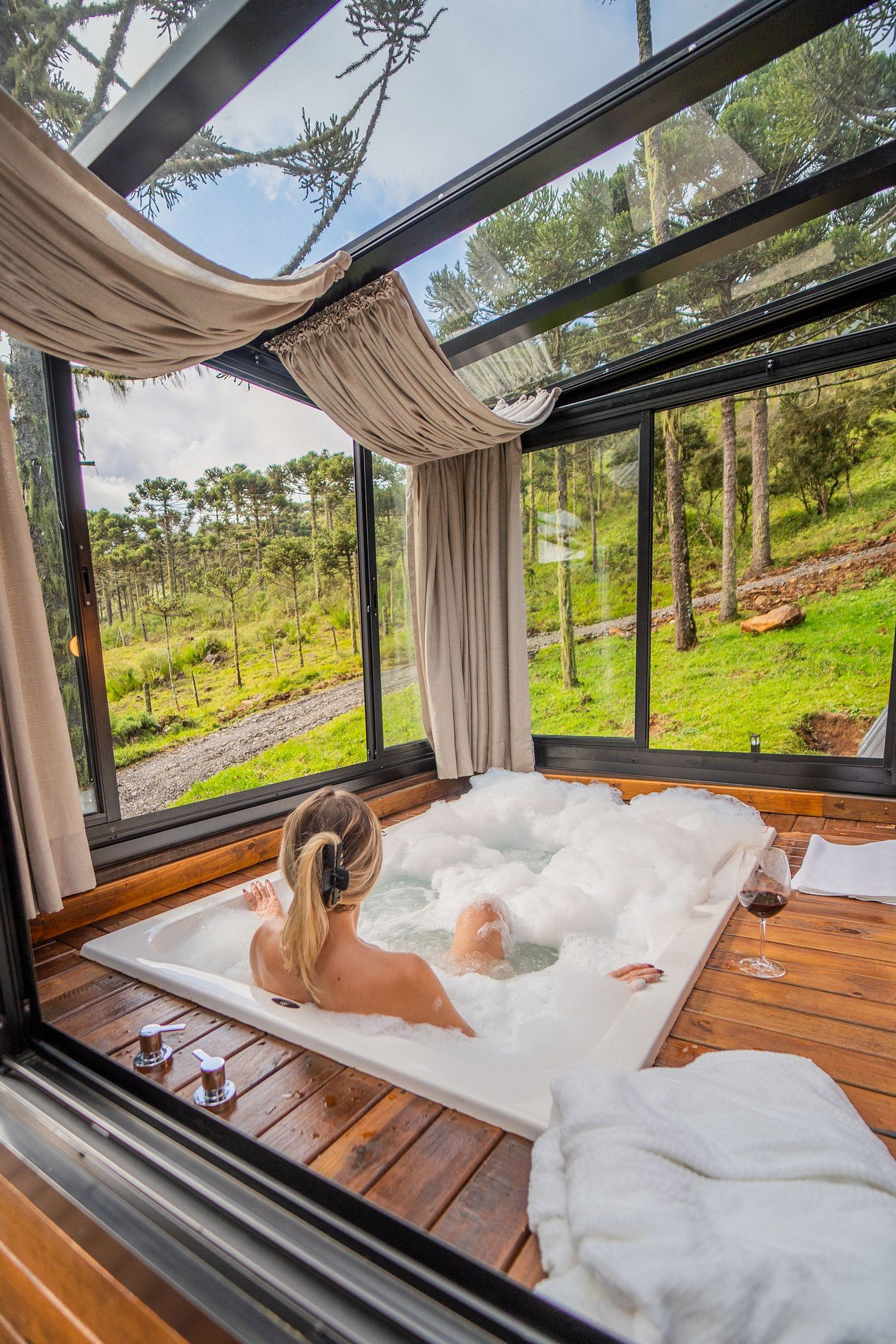 Una habitación con un ventanal grande, una bañera llena de burbujas y suelo de madera.