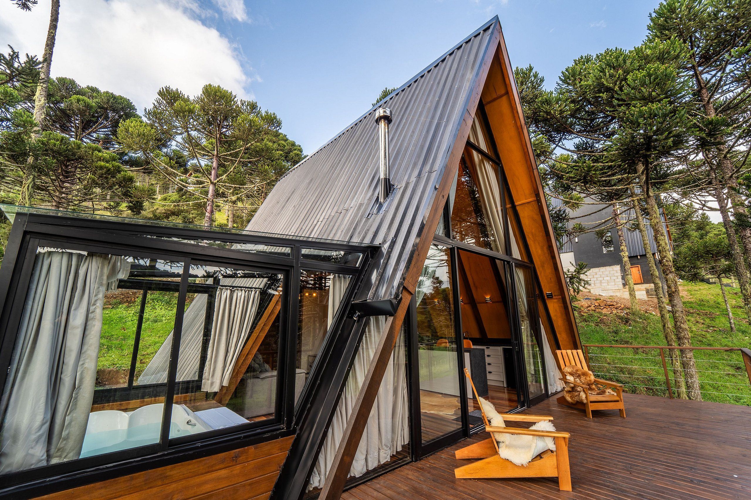 Un chalet moderne en forme de « A », doté d’une terrasse fermée par des baies vitrées, de meubles de terrasse en bois et offrant une vue sur la forêt environnante.