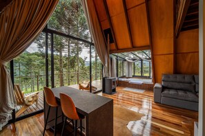 A spacious room with a wooden floor, a sofa, a table with chairs, and large windows offering a view of the forest. - Pousada Refúgio da Boa Vista (Urubici)