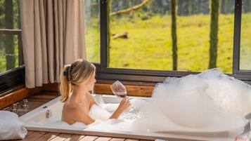 Une femme se détend dans une baignoire, un verre de vin à la main, entourée de lambris en bois et de grandes fenêtres donnant sur un paysage verdoyant.
