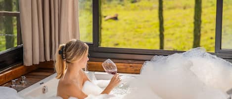 A woman relaxing in a bathtub with a glass of wine, surrounded by wooden paneling and large windows with a view of a green landscape.