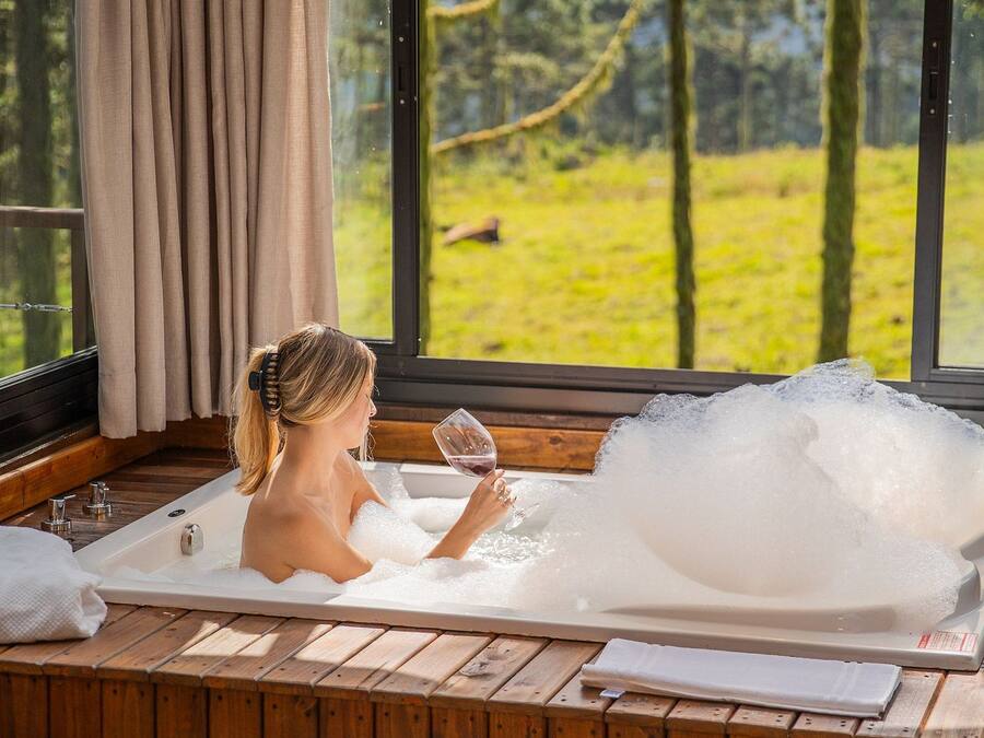 Une femme se détend dans une baignoire, un verre de vin à la main, entourée de lambris en bois et de grandes fenêtres donnant sur un paysage verdoyant.