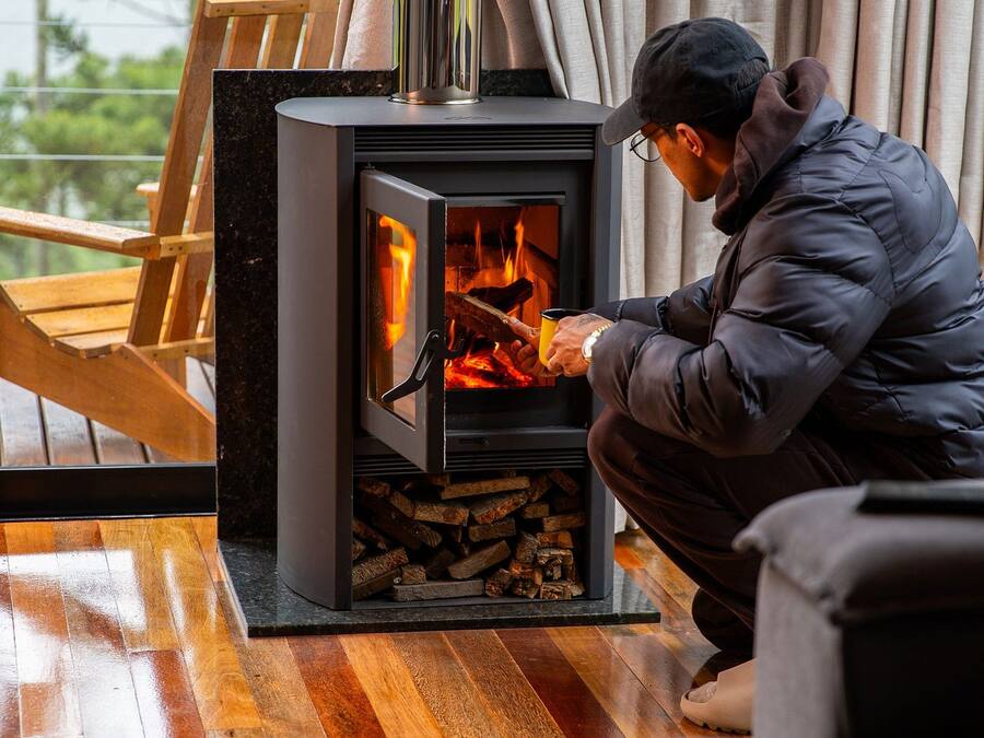 A person sitting by a wood-burning stove in a room with wooden flooring and a view of greenery outside.