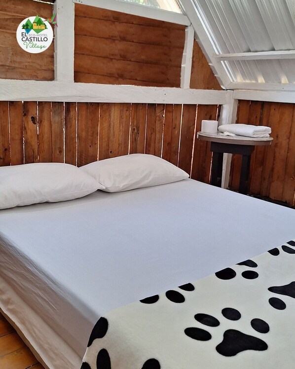 A bed with white bedding and a cow-patterned blanket, wooden walls, and a small table with towels. - ECOHOTEL EL CASTILLO VILLAGE  (Cali)