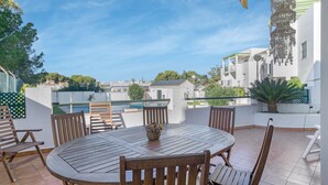 A round wooden table with chairs, a potted plant, and a hanging decorative item on a terrace with a view of buildings and a pool.