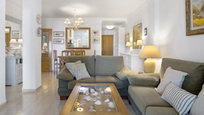 A living room with a sofa, a coffee table with decorative items, a dining area with chairs, and a kitchen in the background.