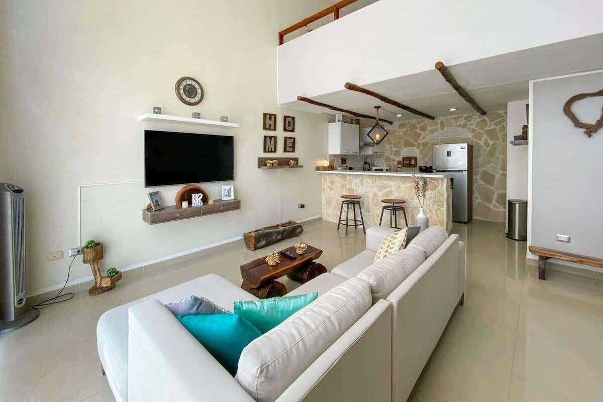 A modern living room with a large sofa, a flat-screen TV, and a kitchen area in the background.