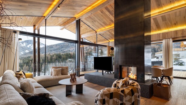 Ein modernes Wohnzimmer mit Sofa, Hocker und Couchtisch, einem Kamin und großen Fenstern mit Blick auf die Berge.