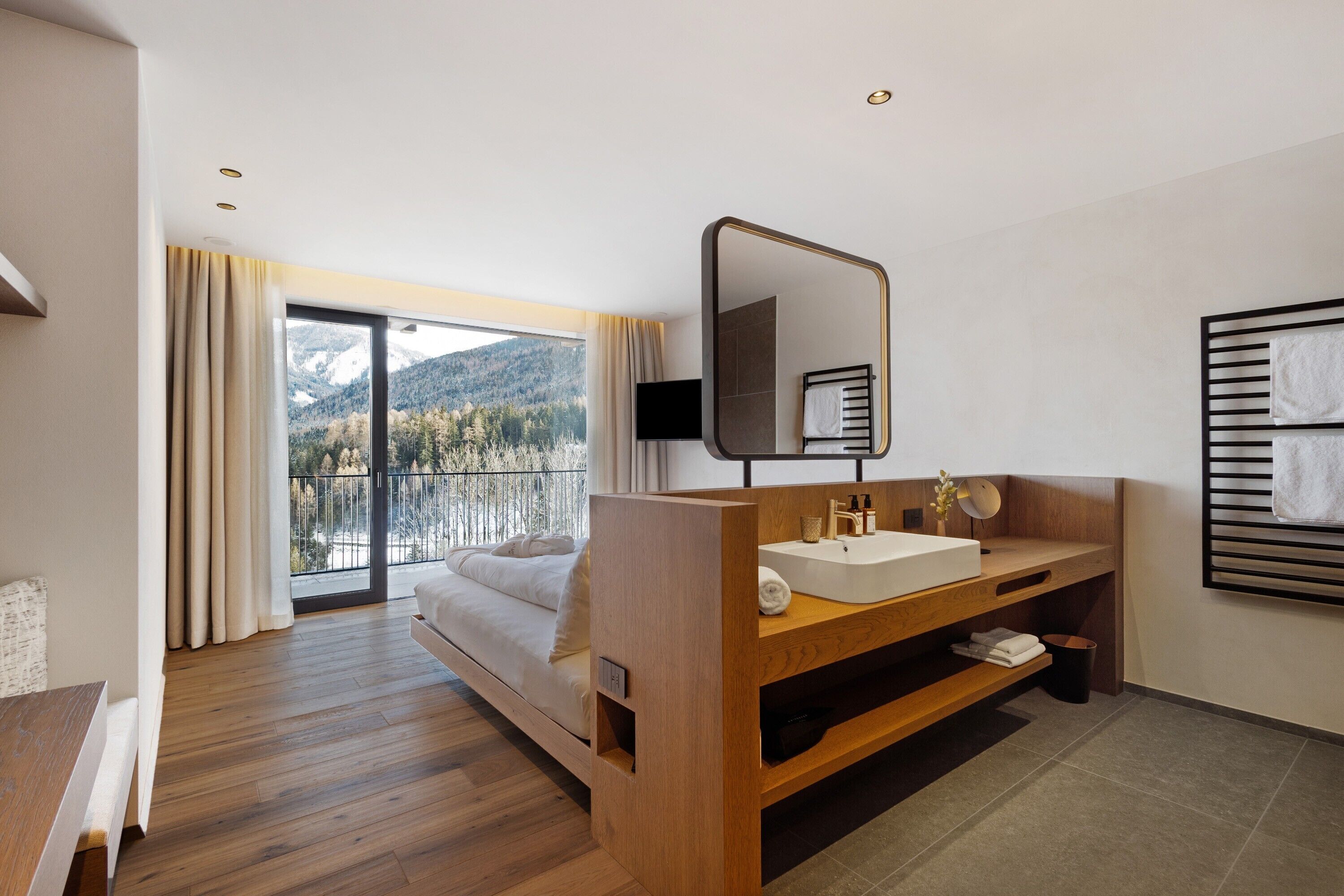 Camera d'albergo con un letto di legno, un'ampia finestra vista montagne, una toeletta in legno con uno specchio e un porta asciugamani nero.