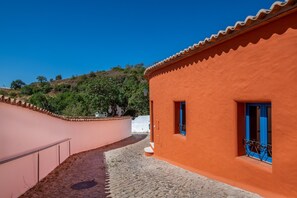 Uma estreita viela de paralelepípedos, com um edifício à direita que apresenta fachada vermelha, porta azul e uma pequena varanda com grades de metal.