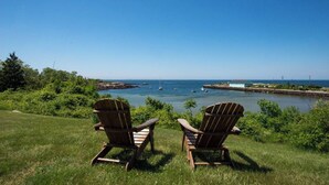Två trästolar i Adirondack-stil, placerade så att de vetter mot havet, med båtar och en byggnad synlig på avstånd.