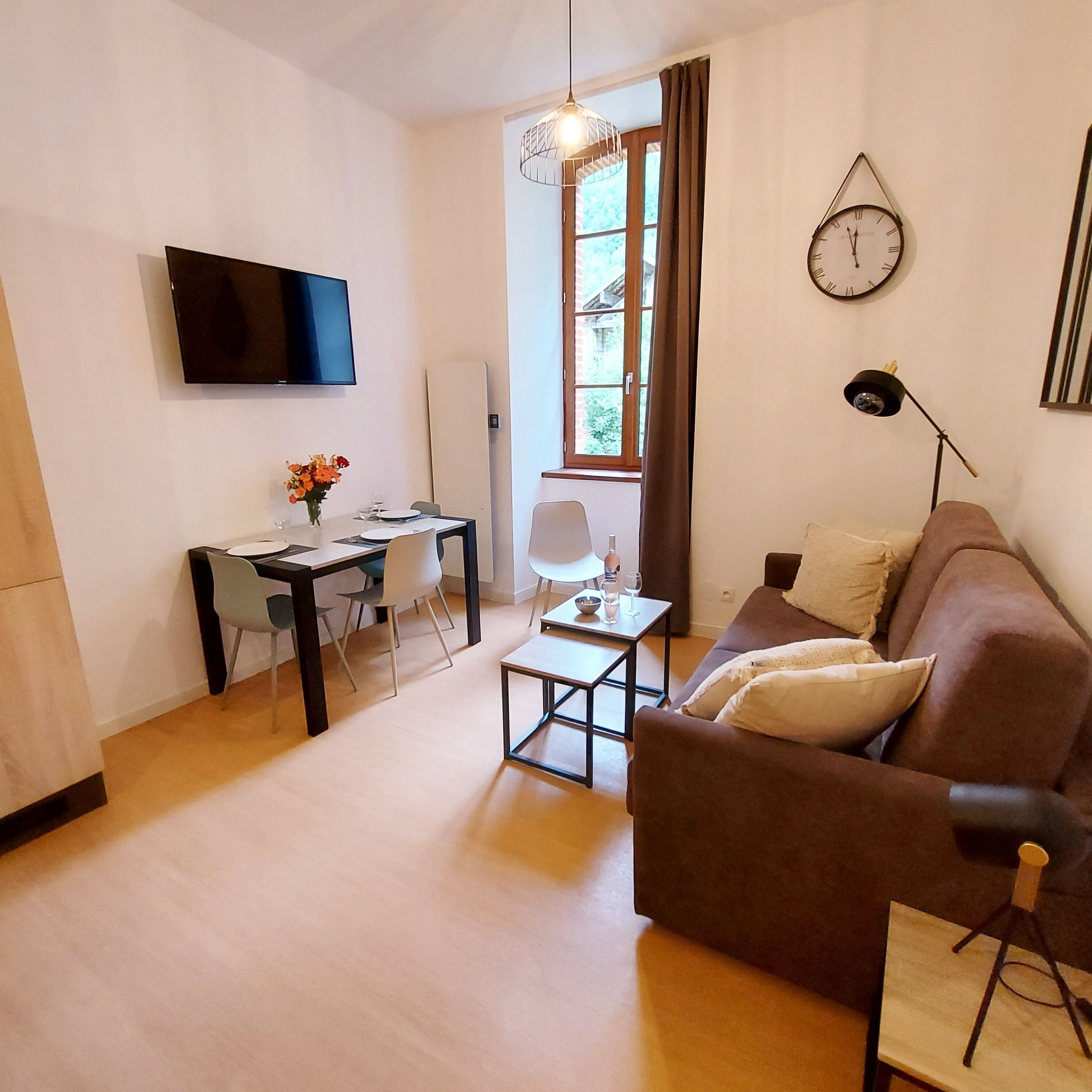 Un salon moderne avec un canapé marron, une table à manger avec des chaises, un téléviseur fixé au mur et une fenêtre avec des rideaux.