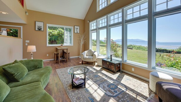 A spacious living room with a large window, a green couch, a glass coffee table, and a view of the outdoors. - Waterfront Bellingham Bliss: Steps to Beach! (Bellingham)