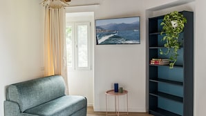 A living room with a grey sofa, a television mounted on the wall displaying a coastal scene, a small round table with a candle, and a bookshelf with green plants.