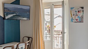 A room with a TV displaying a coastal scene, a balcony with a view of buildings and flowers, and a piece of abstract art.