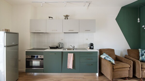 A compact kitchen with white cabinets, a built-in oven, and a sink. There is a brown armchair with a cushion and a small table with a kettle and a coffee maker.