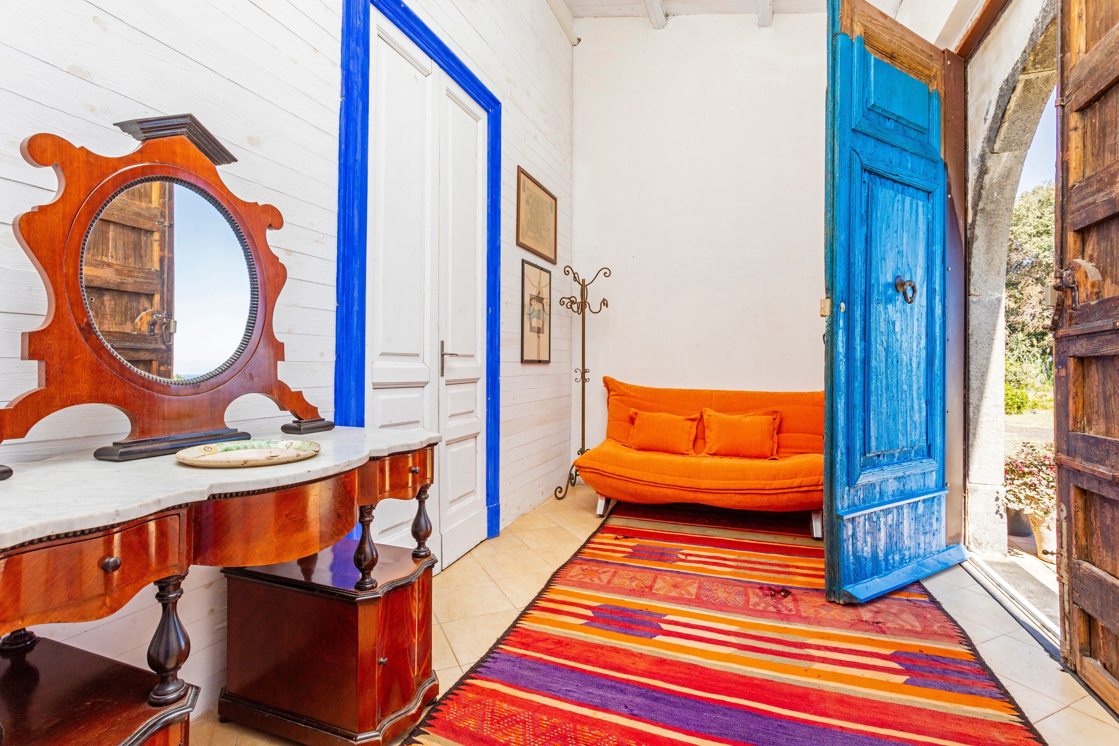 Ein Zimmer mit einem hölzernen Schminktisch, einem farbenfrohen Streifen Teppich, einem orangefarbenen Sofa und einer blauen Tür.