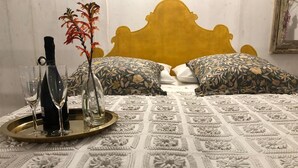 Un lit recouvert d’une courtepointe à motifs, deux oreillers ornés de motifs floraux, un plateau avec deux verres et une bouteille, et un vase rempli de fleurs rouges.