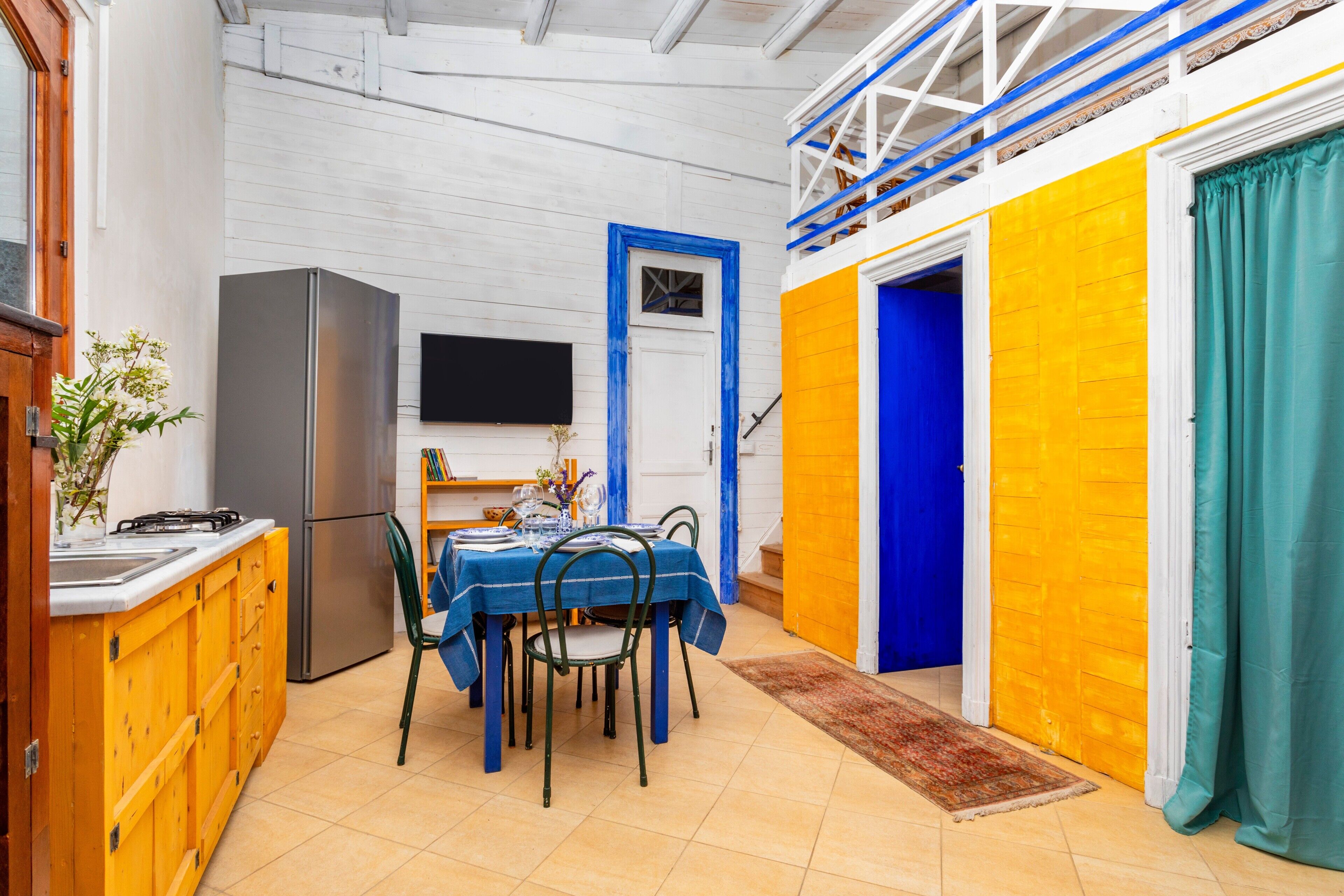 A kitchen with a dining table, chairs, a refrigerator, and a TV on the wall.
