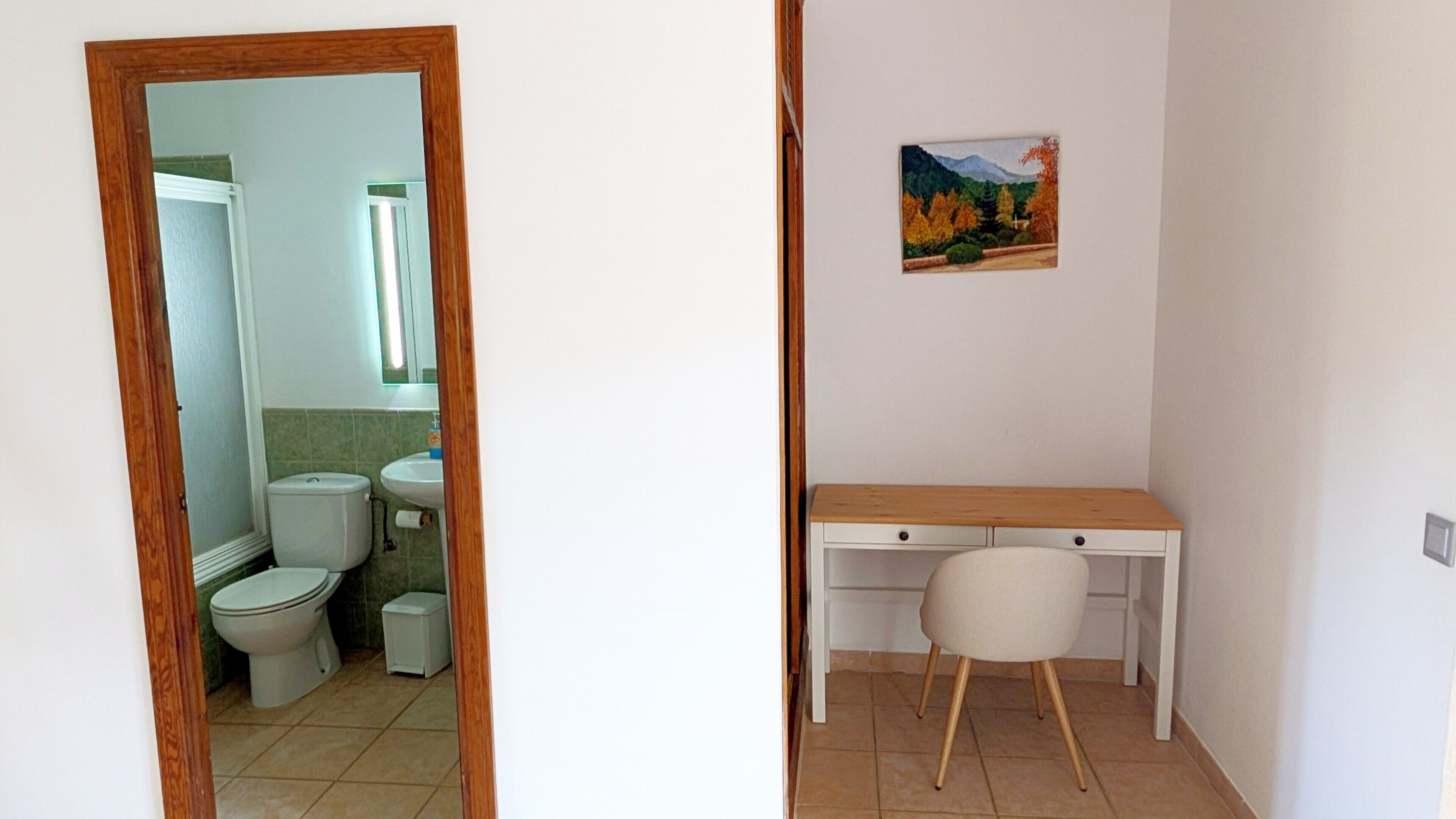 Een badkamer met toilet, wastafel en spiegel, en een bureau met stoel in een aparte kamer.