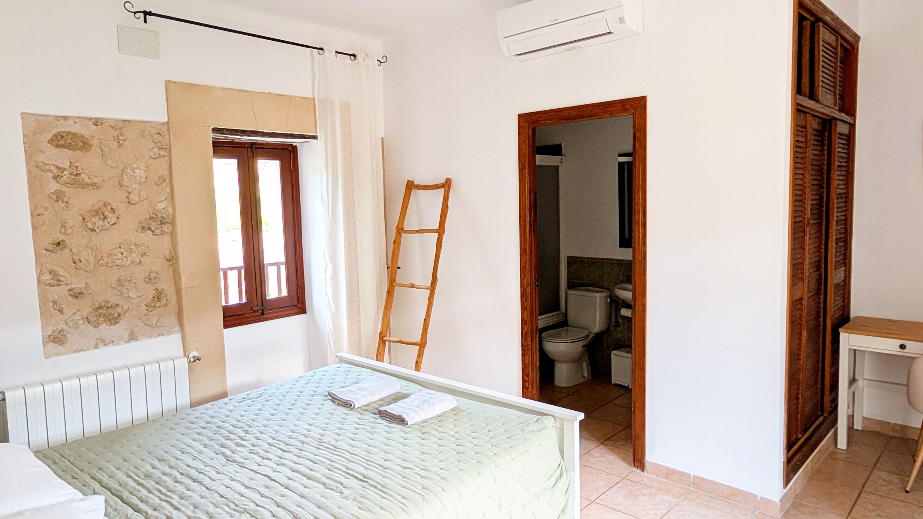 Een slaapkamer met een bed, een raam, een radiator, een airconditioner en een badkamer zichtbaar door een open deur.
