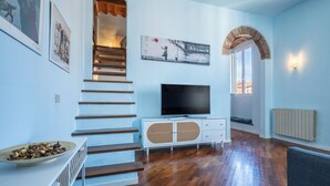 A living room with a wooden staircase, a television, and a white cabinet with a basket. - Libeccio Urban Suite by Pleiades Home (Livorno)
