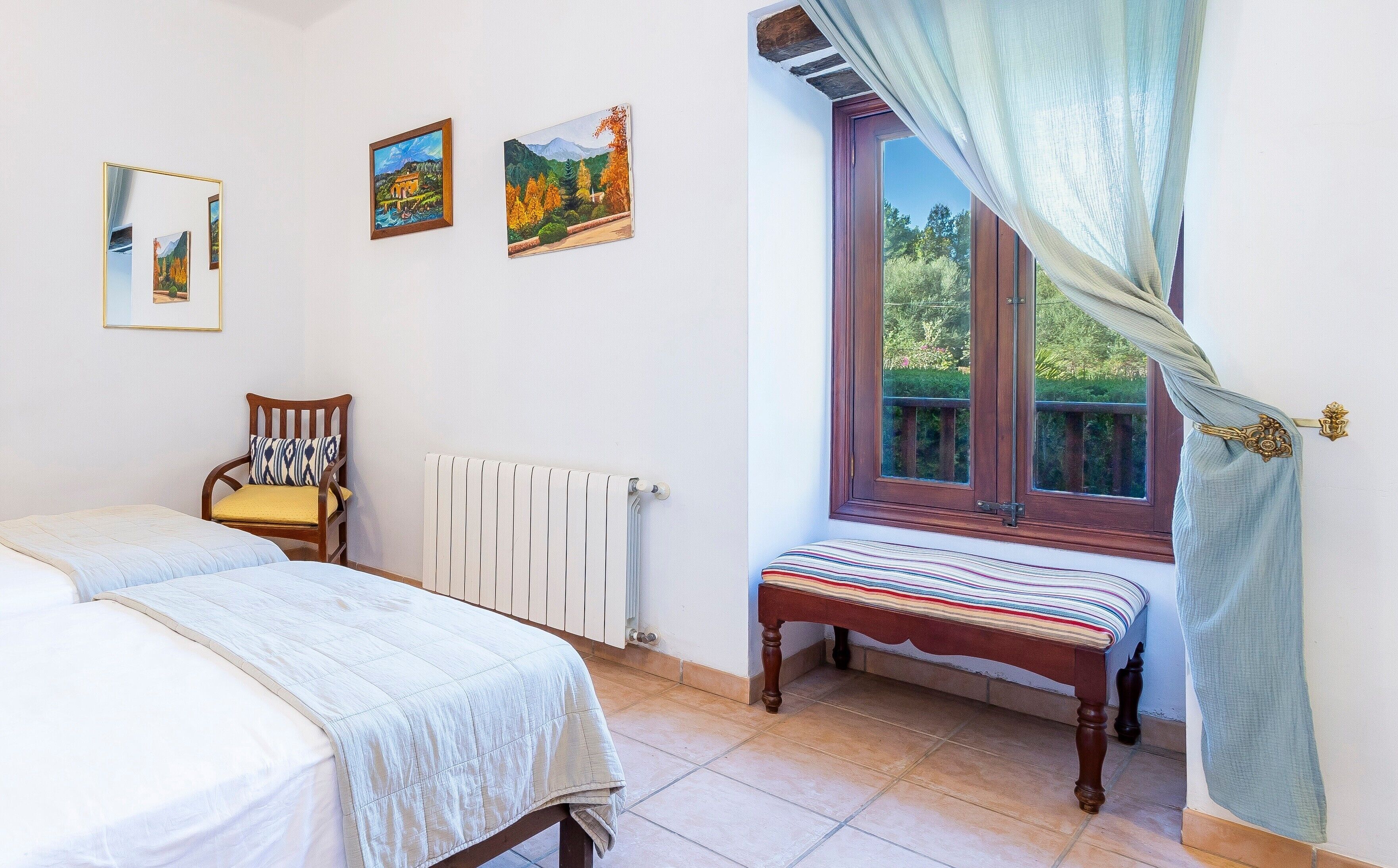 A bedroom with a bed, a chair, a bench, a radiator, and a window with curtains.