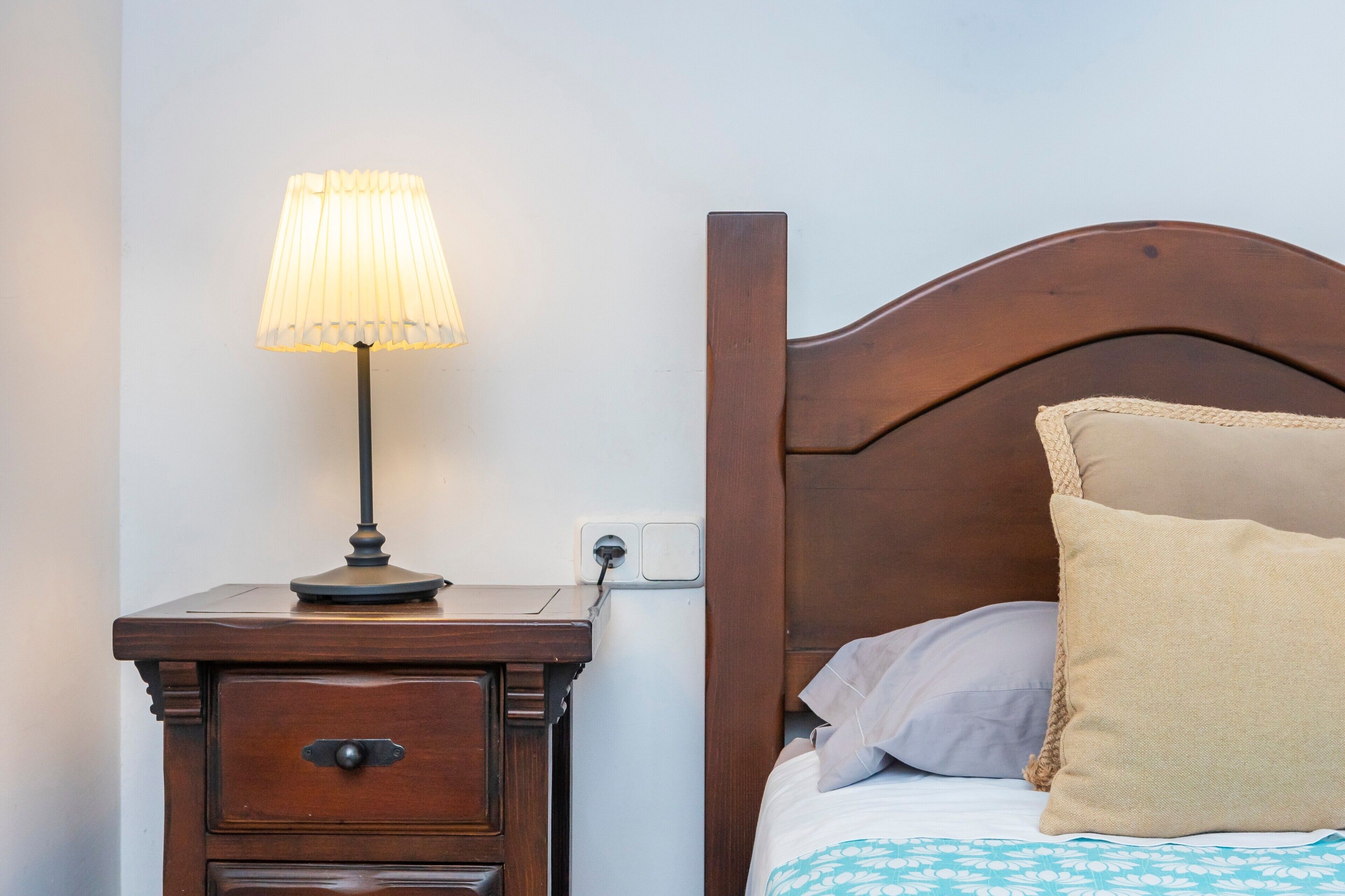 Una mesita de noche de madera con una lámpara, una cama con cabecera y una sola almohada.