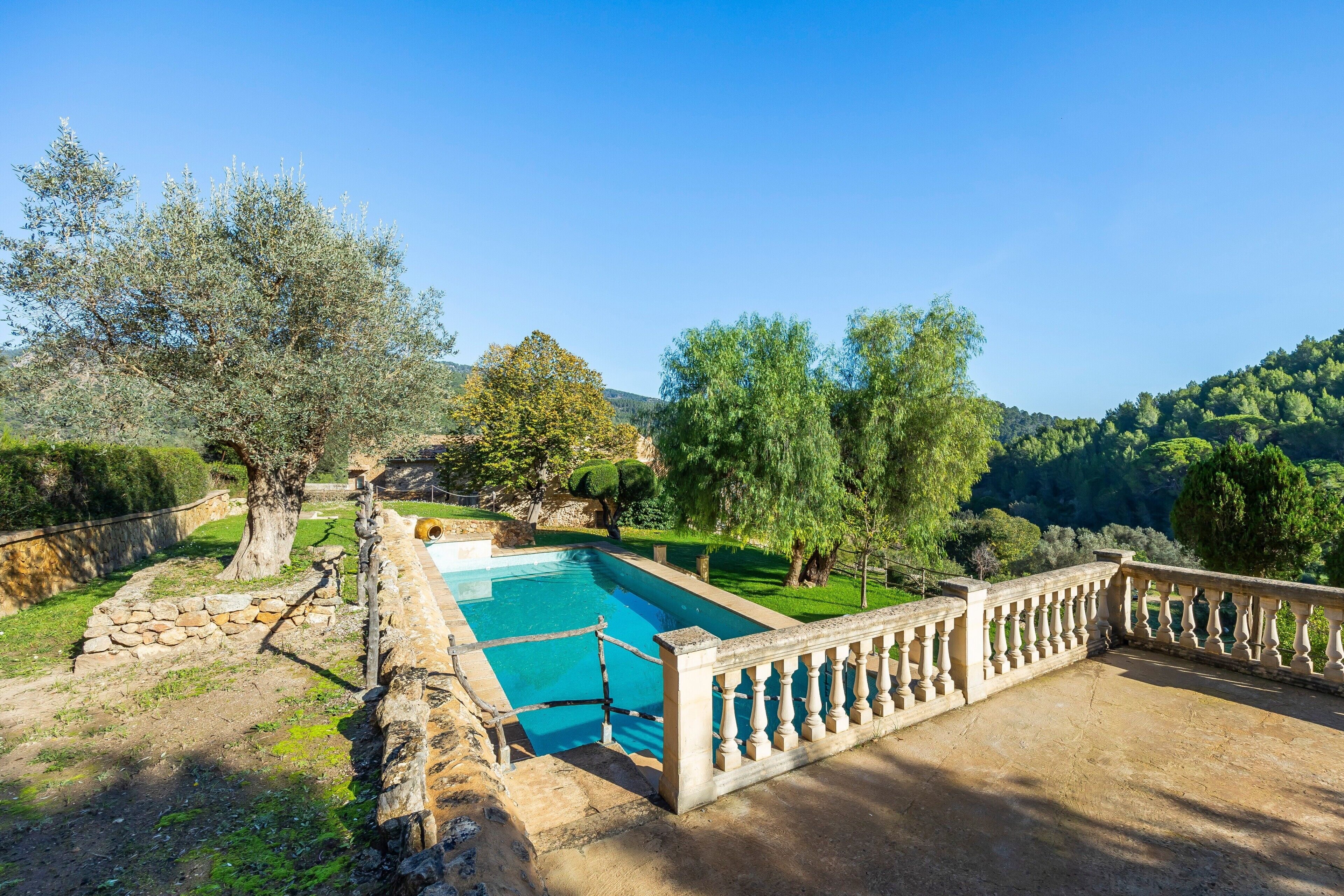 A pool with a stone wall and a balustrade, surrounded by trees and a hilly landscape.