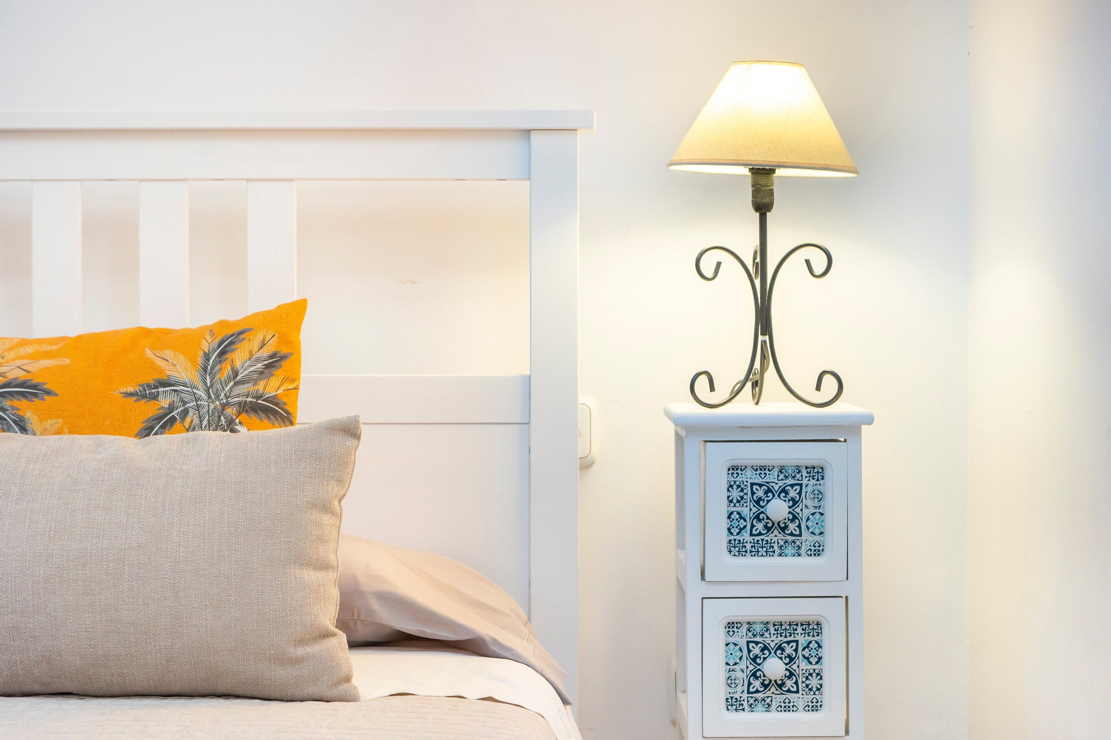 A bed with a white headboard, a yellow pillow, a beige pillow, and a bedside table with a lamp and two drawers.