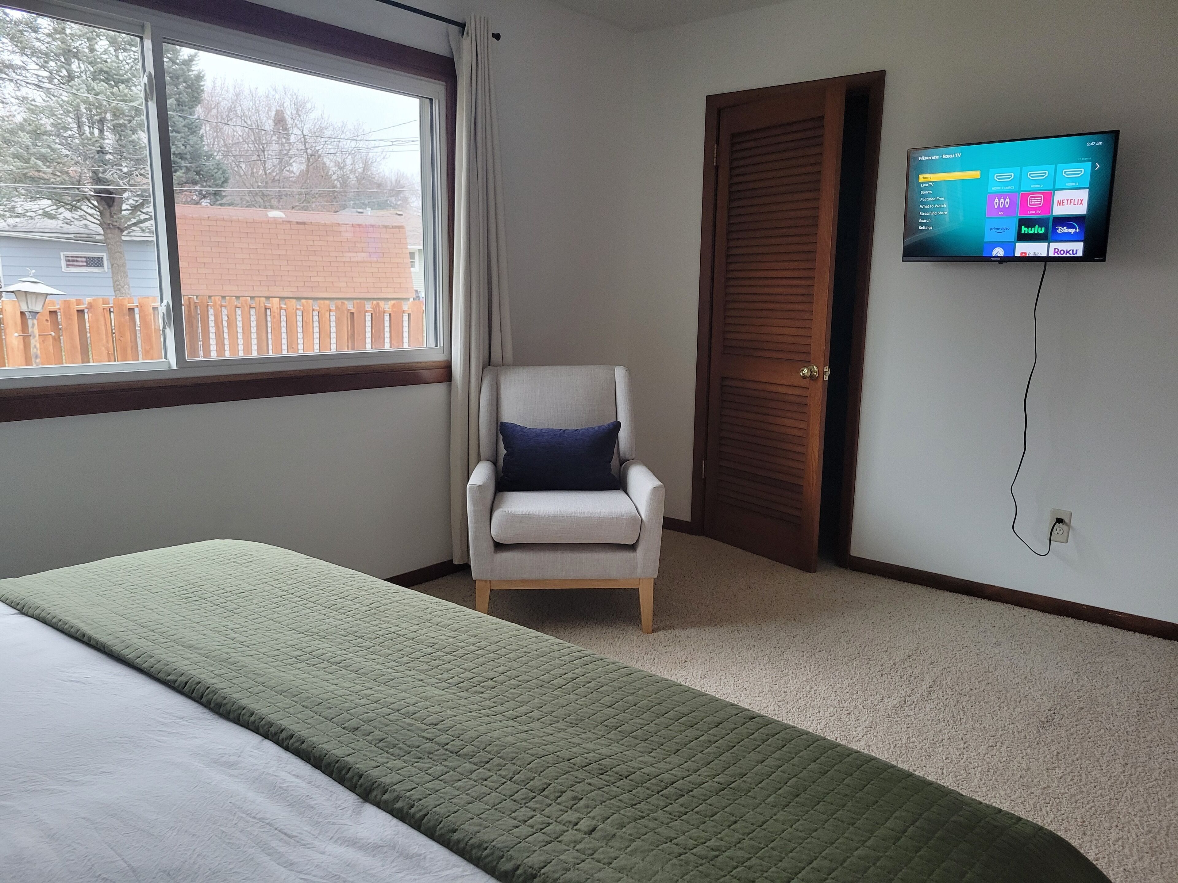 Ein Hotelzimmer mit Bett, Sessel, an der Wand montiertem Fernseher und Fenster mit Blick auf einen Zaun und Gebäude.