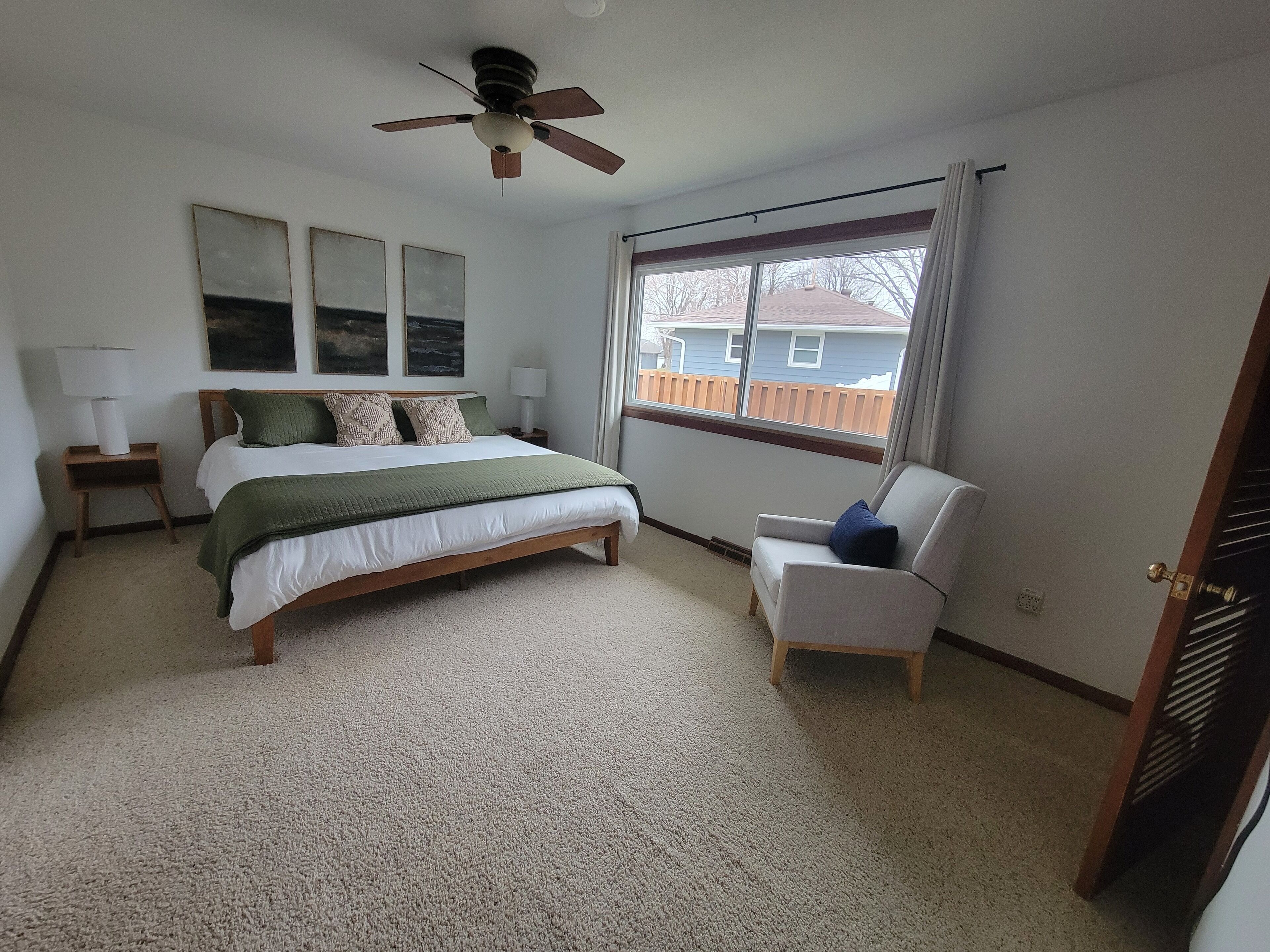 Une chambre à coucher comprenant un lit, une chaise, un ventilateur de plafond et une fenêtre donnant sur une maison.