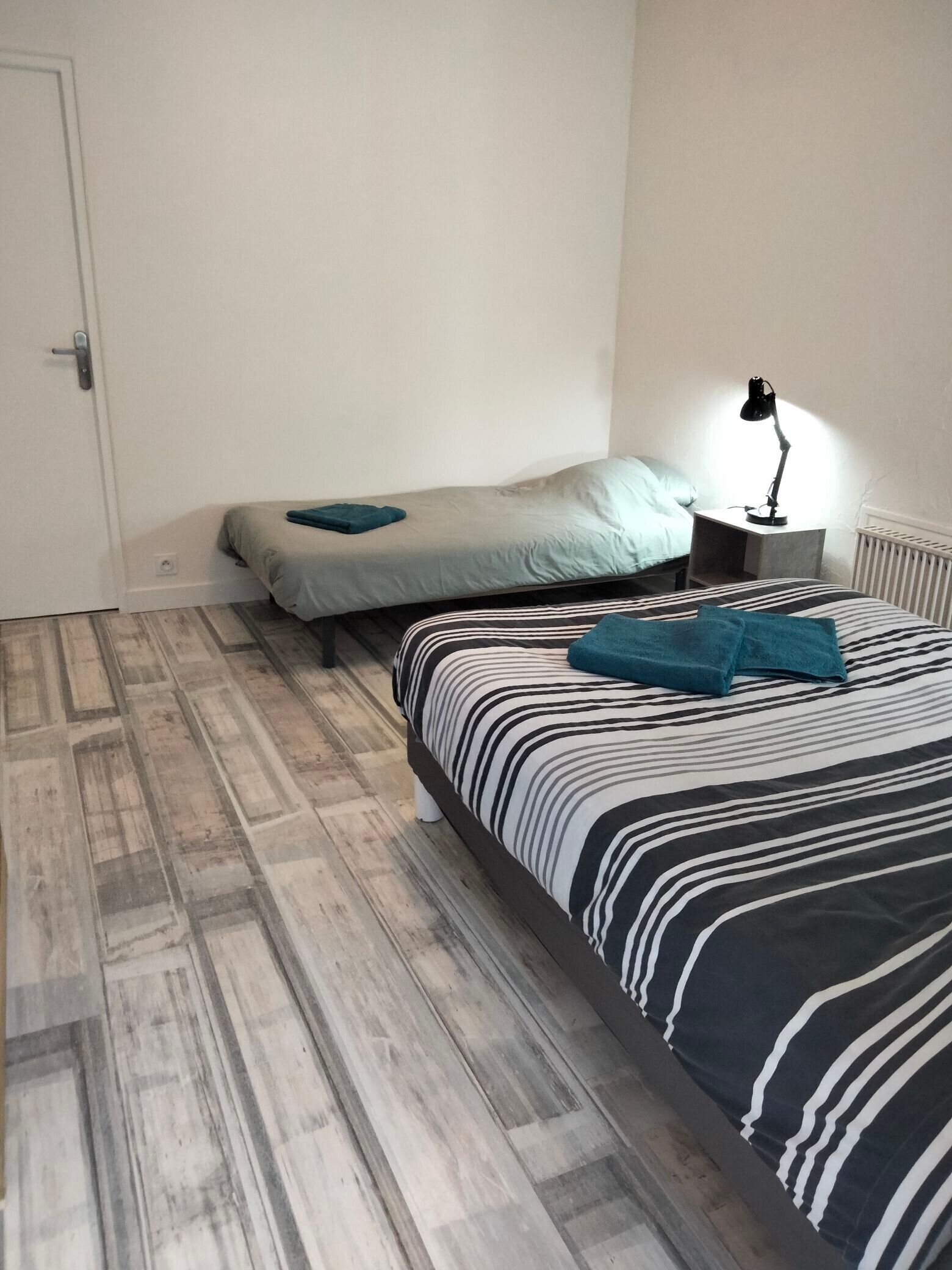 A bedroom with two beds, a nightstand with a lamp, and a radiator.